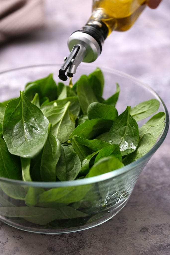 olive oil being poured on top of spinach leaves