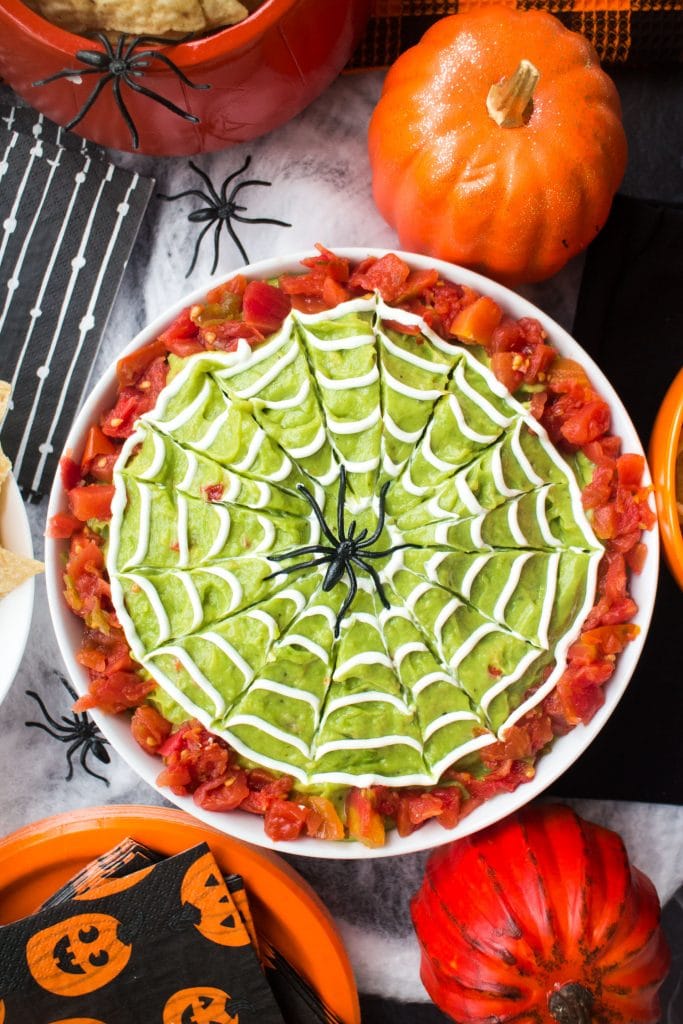 halloween guacamole dip in white bowl with sour cream spiderwebs on top on halloween decorated table