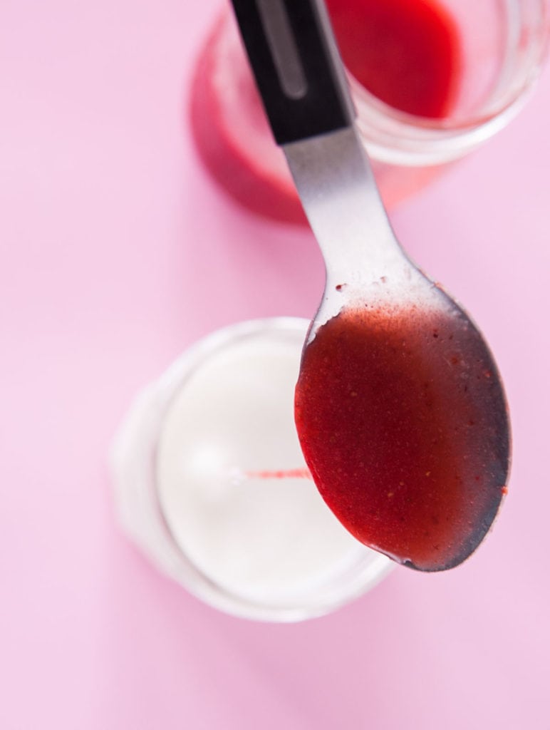 spoon filled with strawberry syrup going into a cup of milk.