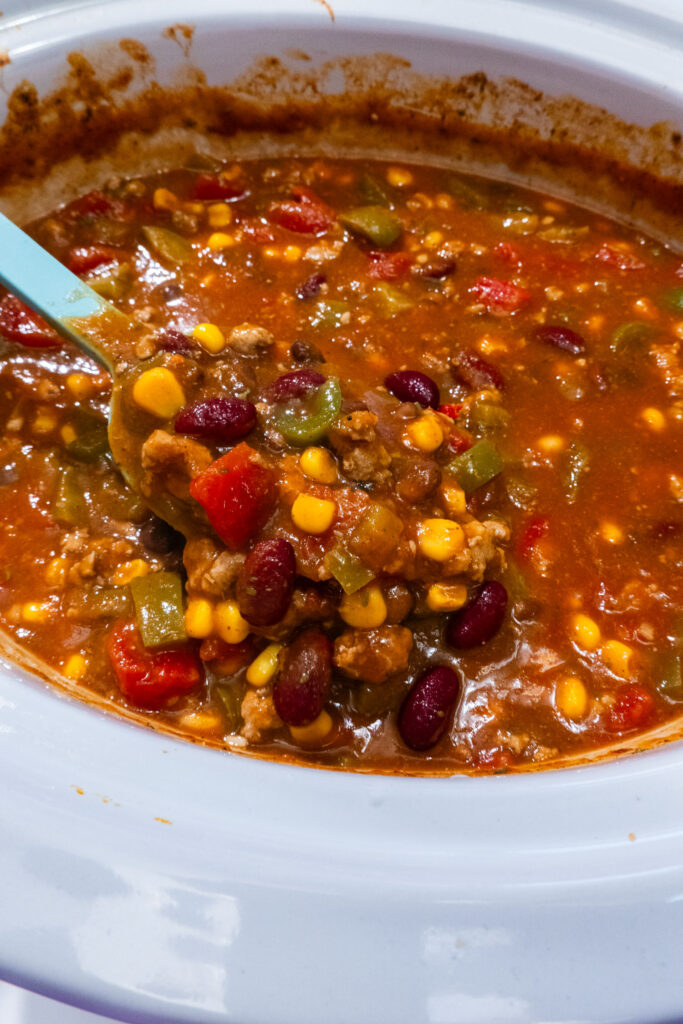 A blue ladle scoops chili containing ground meat, kidney beans, corn, and bell peppers from a slow cooker.