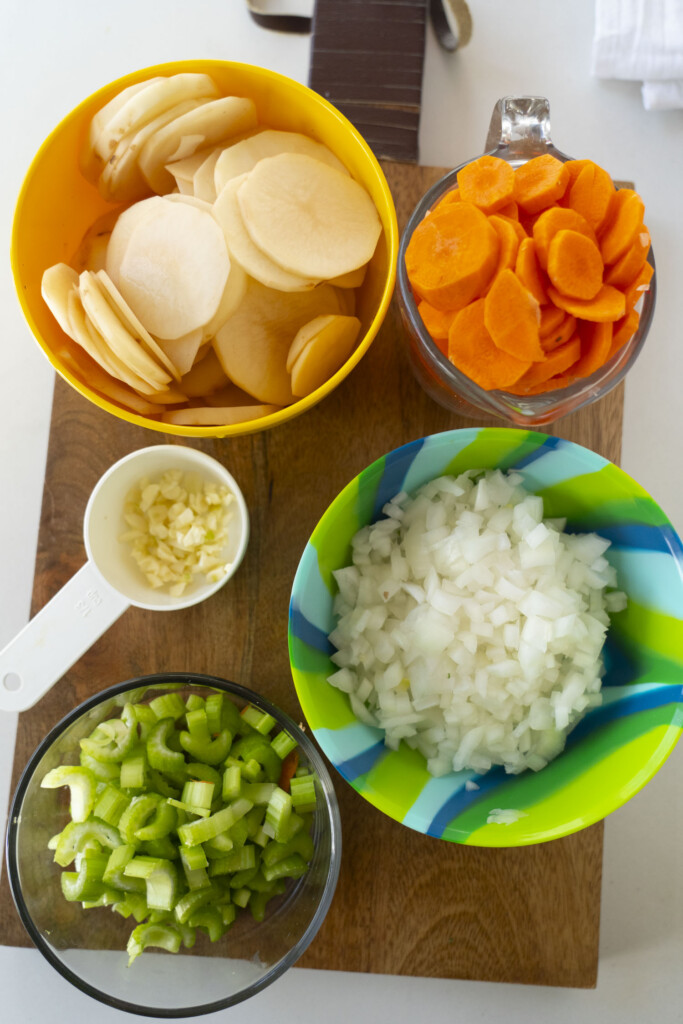 A wooden board holds bowls of chopped vegetables: sliced potatoes in a yellow bowl, sliced carrots in a clear cup, diced onions in a colorful bowl, chopped celery in a glass bowl, and minced garlic in a white measuring cup.