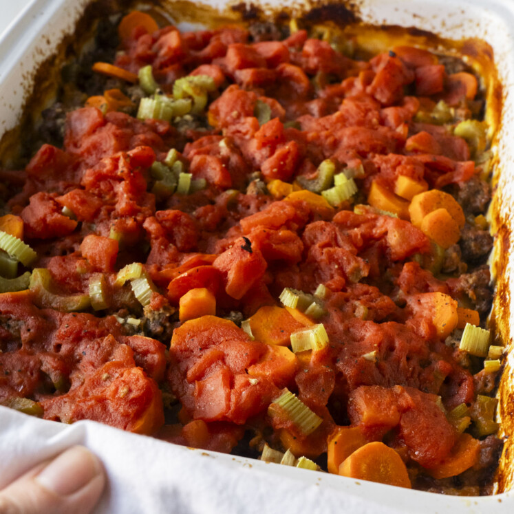 Close-up of a baked casserole in a white dish, topped with chunks of tomato, sliced carrots, and celery. A hand is holding the edge of the dish, ready to serve.