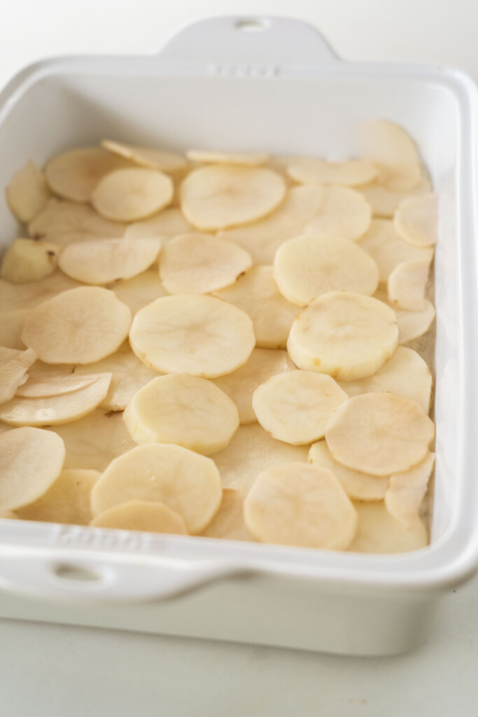 Thinly sliced raw potatoes are arranged in a single layer in a white rectangular baking dish, ready to be cooked.