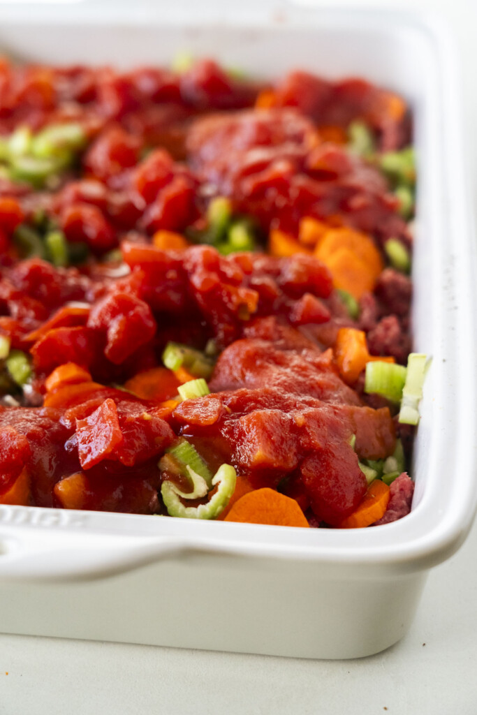 A close-up of a white baking dish filled with chopped celery, carrots, ground meat, and topped with chunky tomato sauce, ready to be cooked.