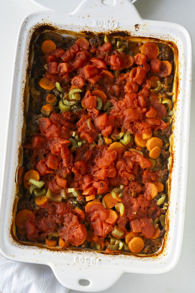 A white casserole dish filled with a baked mixture of chopped carrots, celery, ground meat, and diced tomatoes, all layered and cooked together. The edges of the dish are lightly browned.