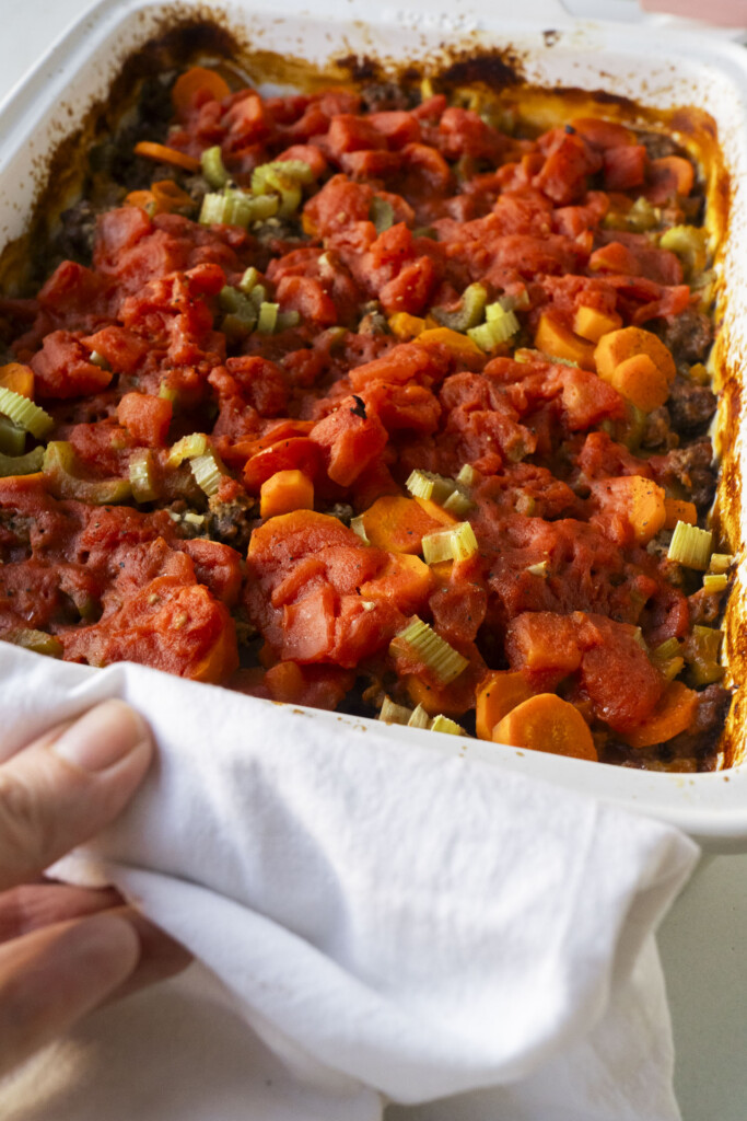 A hand holding a white towel grips the edge of a casserole dish filled with baked vegetables, including carrots, celery, and tomato sauce.