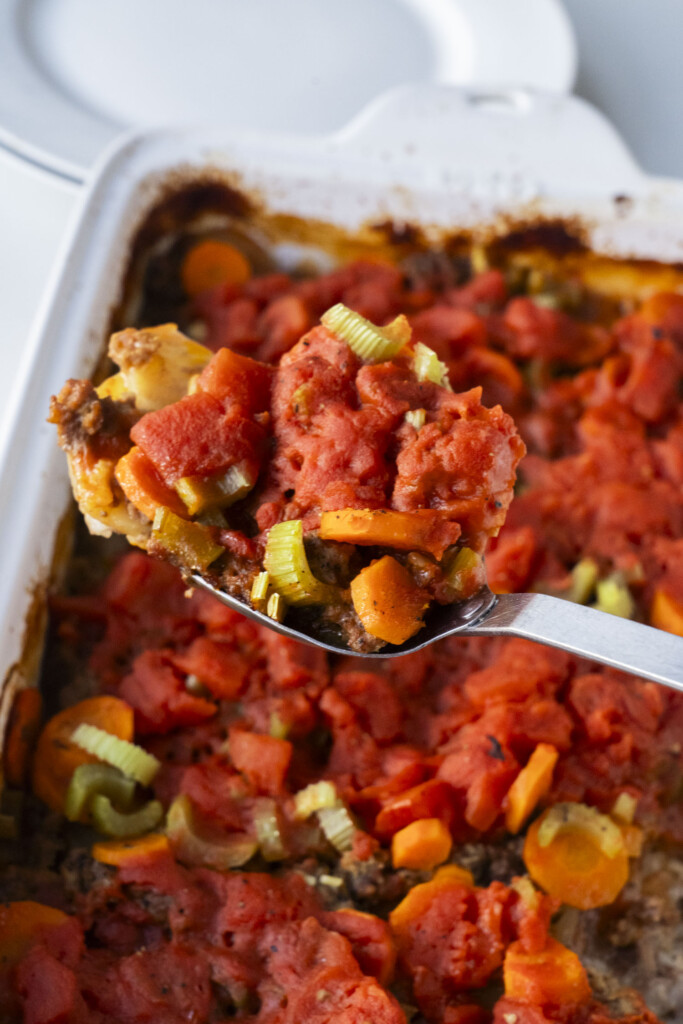 A close-up of a spoon lifting a serving of baked casserole with chunks of tomato, carrots, celery, and other vegetables from a white baking dish.