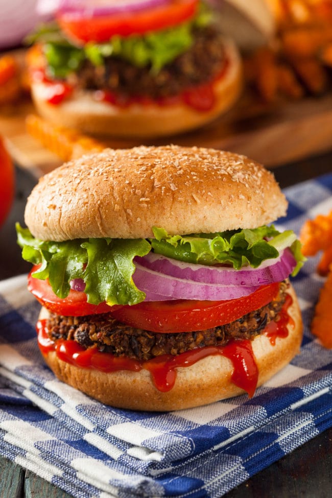 Veggie burger patty in a brown bun along with lettuce, tomato and red onion. Ketchup is dripping out of the bun and the whole burger is on a blue checkered napkin.