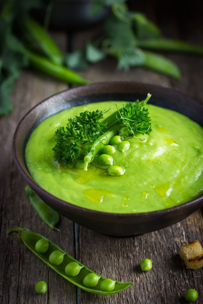 creamy pea soup in brown wooden bowl with fresh peas surrounding it