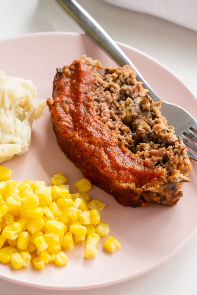 cheesy meatloaf on pink plate with tomato sauce glaze with mashed potatoes and corn
