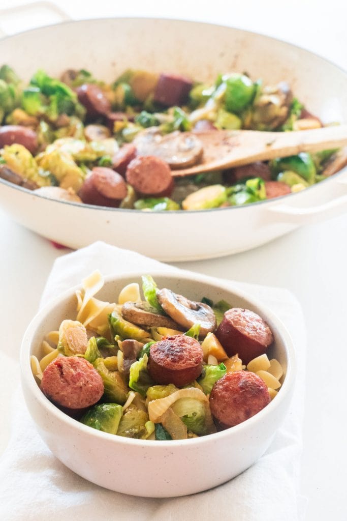 bowl of egg noodles with kielbasa and brussels sprouts with skillet behind it