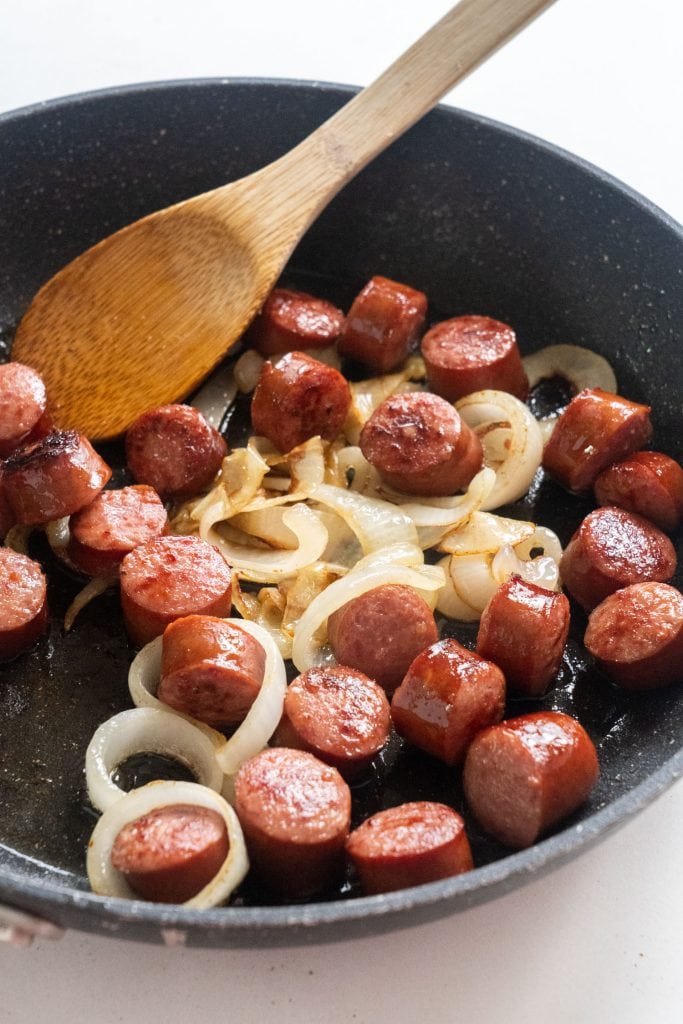 chopped up sausage with fried onions in skillet with wooden spoon