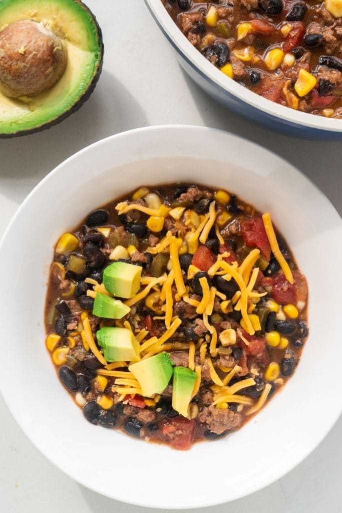 taco soup in white bowl with shredded cheese and avocado on top