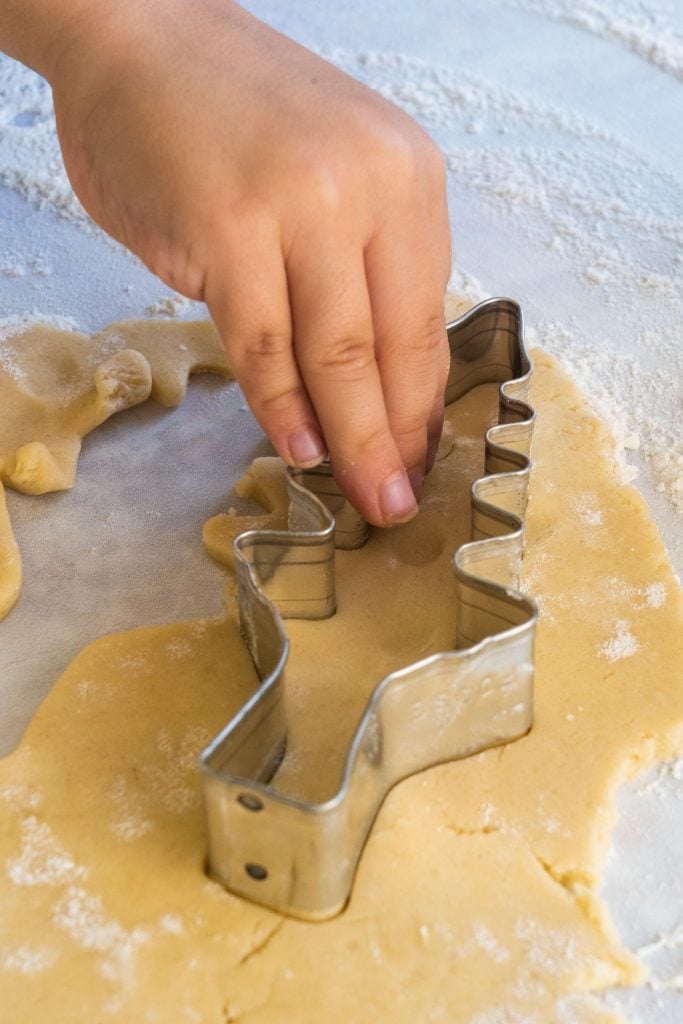 kids hand using tree cookie cutter to cut out sugar cookie dough on floured surface