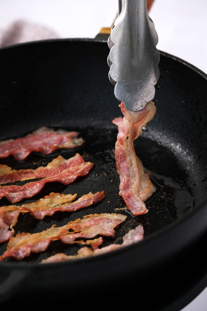 tongs picking up bacon in frying pan that's brown on both sides.
