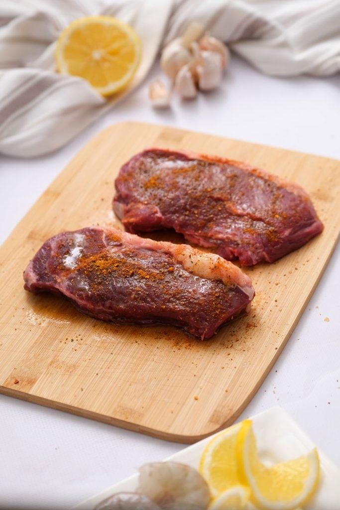 steak seasoning and oil on new york strip steaks on cutting board
