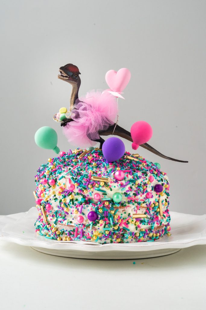 3 layer cake with purple, teal and pink sprinkles with tutu wearing dinosaur on top.