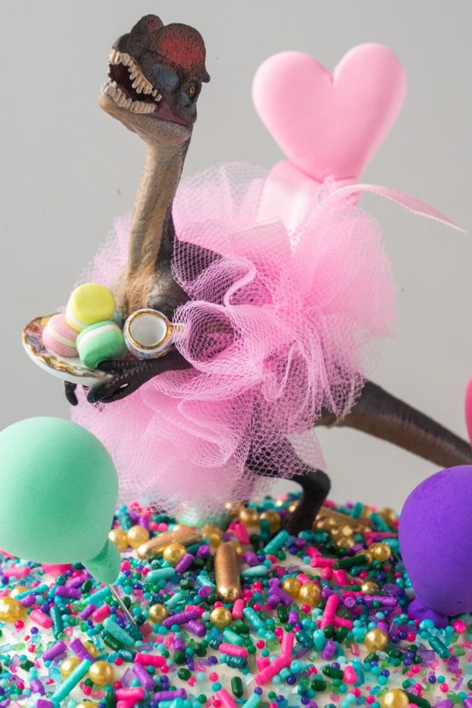 dinosaur wearing tutu on top of cake holding a plate of macarons