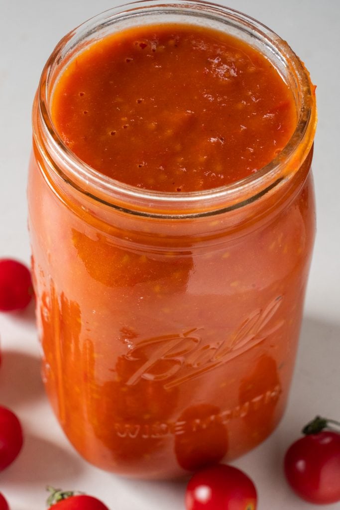 mason jar filled with tomato sauce with cherry tomatoes surrounding it on table.