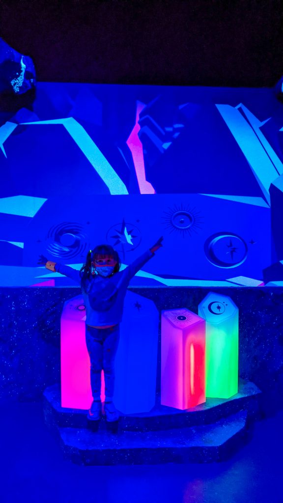 girl raising her arms in front of crystal installation that is lit up rainbow colors.