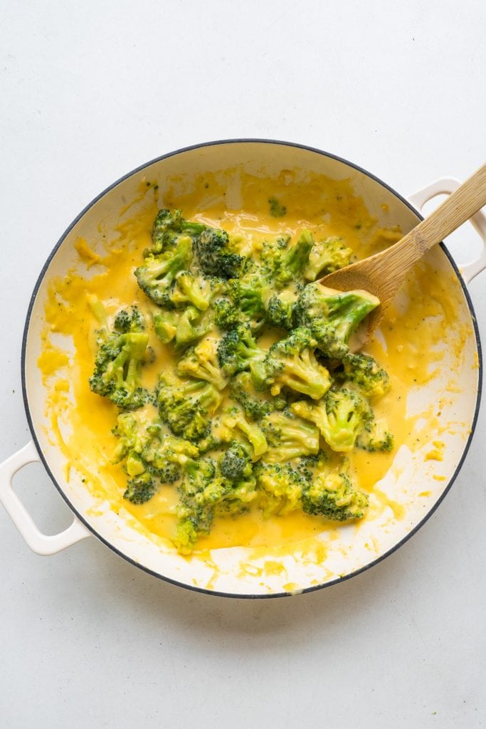 spoon stirring broccoli and cheese in white pan on table.