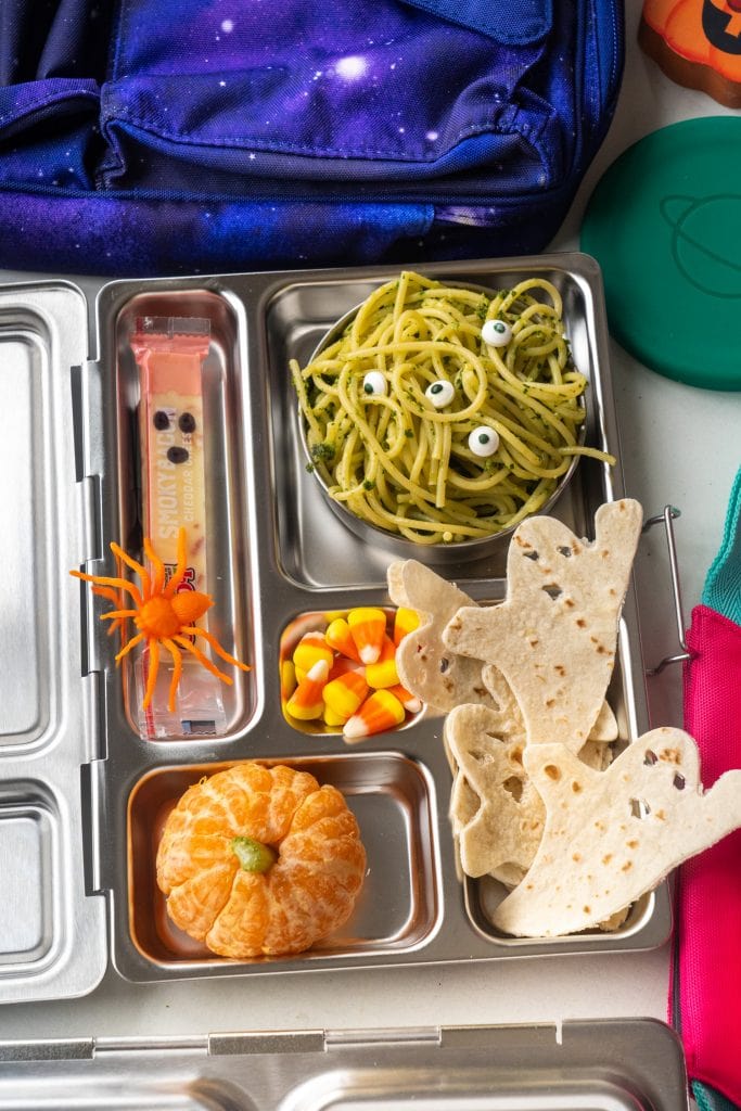 lunch box filled with pasta, oranges, tortillas, cheese stick and candy, decorated for halloween.