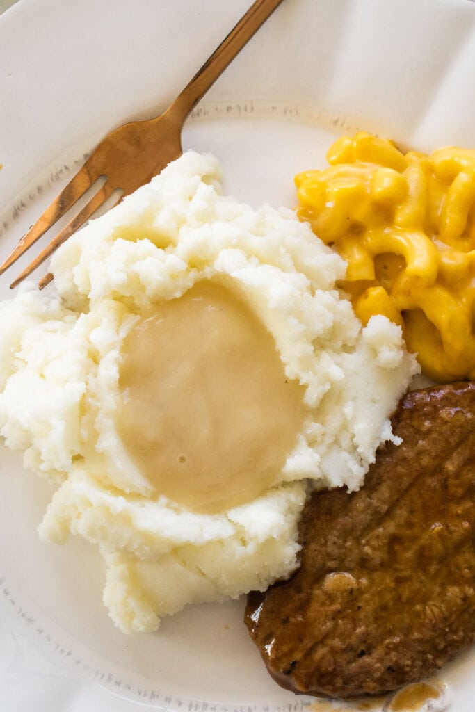 mashed potatoes with gravy on top next to salisbury steak and mac and cheese on plate.