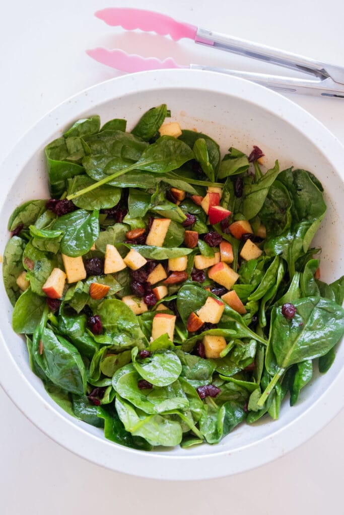 spinach salad with apples and cranberries in white bowl on table.