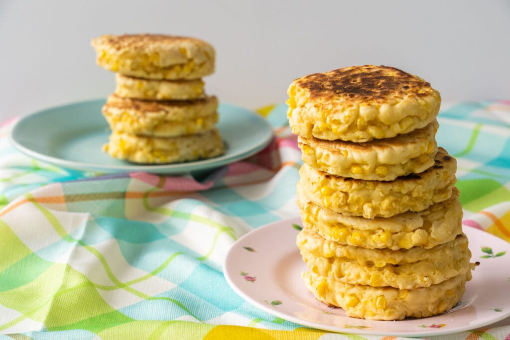 2 large stacks of corn pancakes on plate on plaid tablecloth.