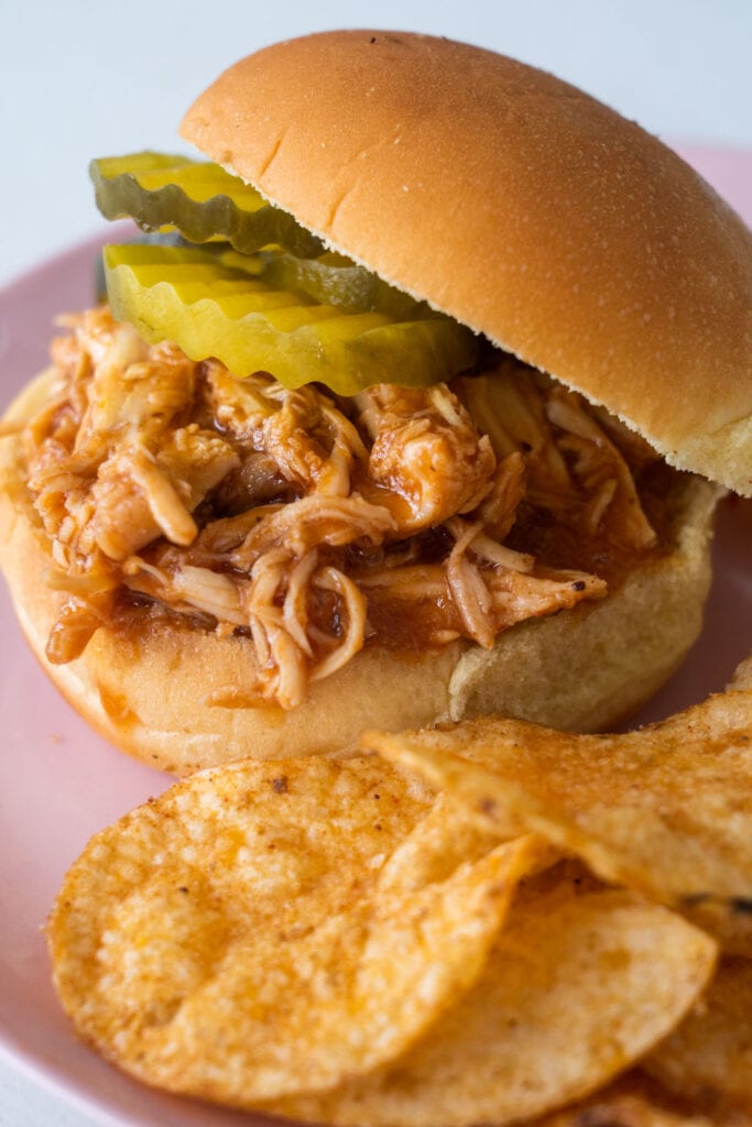 closeup of bbq chicken sandwich with pickles on on plate next to bbq chips.