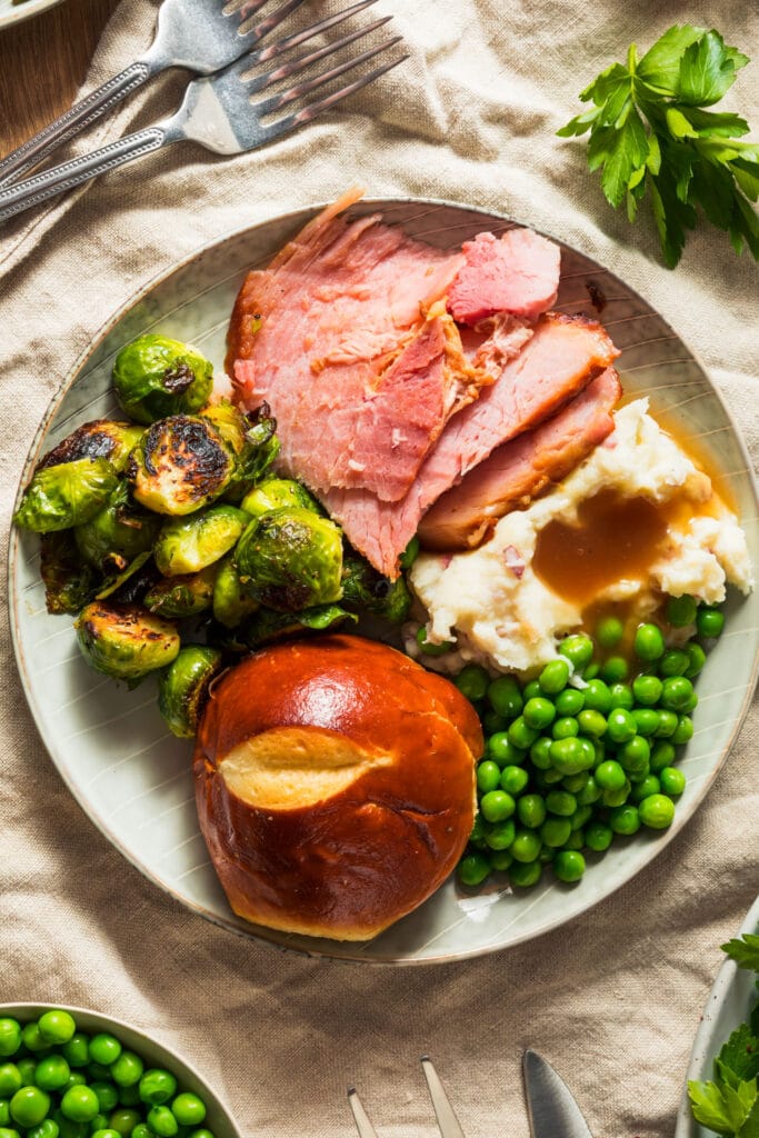 ham on plate with Brussels sprouts, mashed potatoes, peas and roll.
