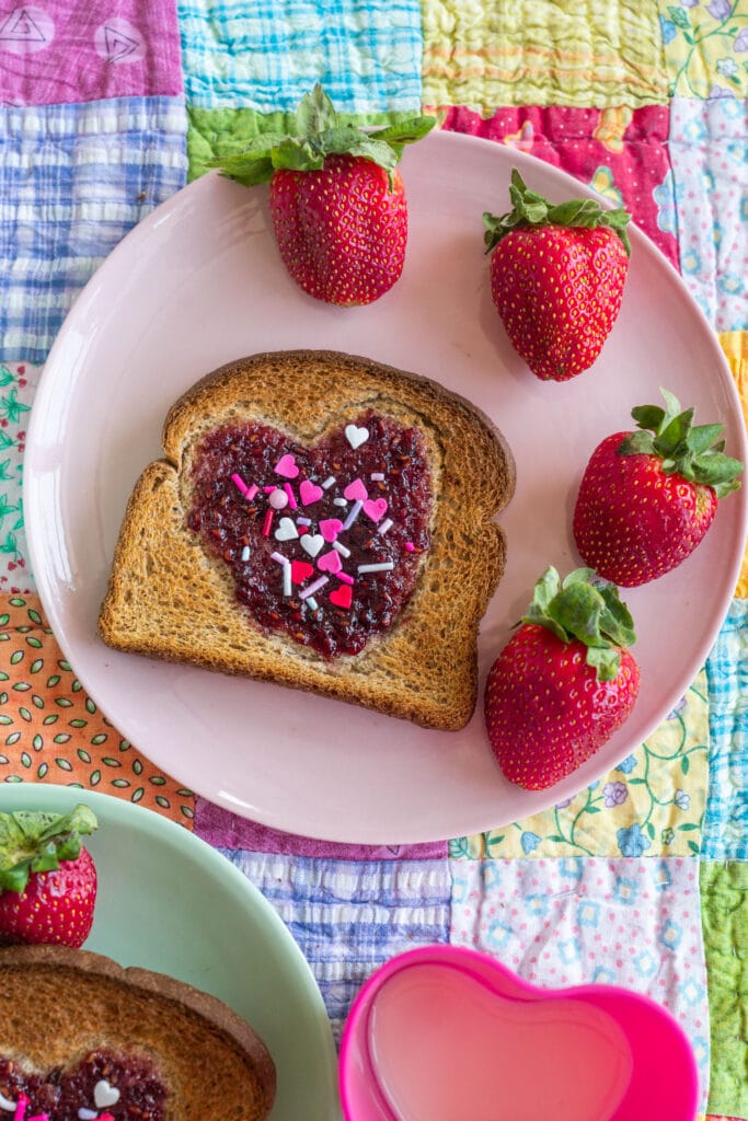 heart toast with jam next to strawberries served on pink plate