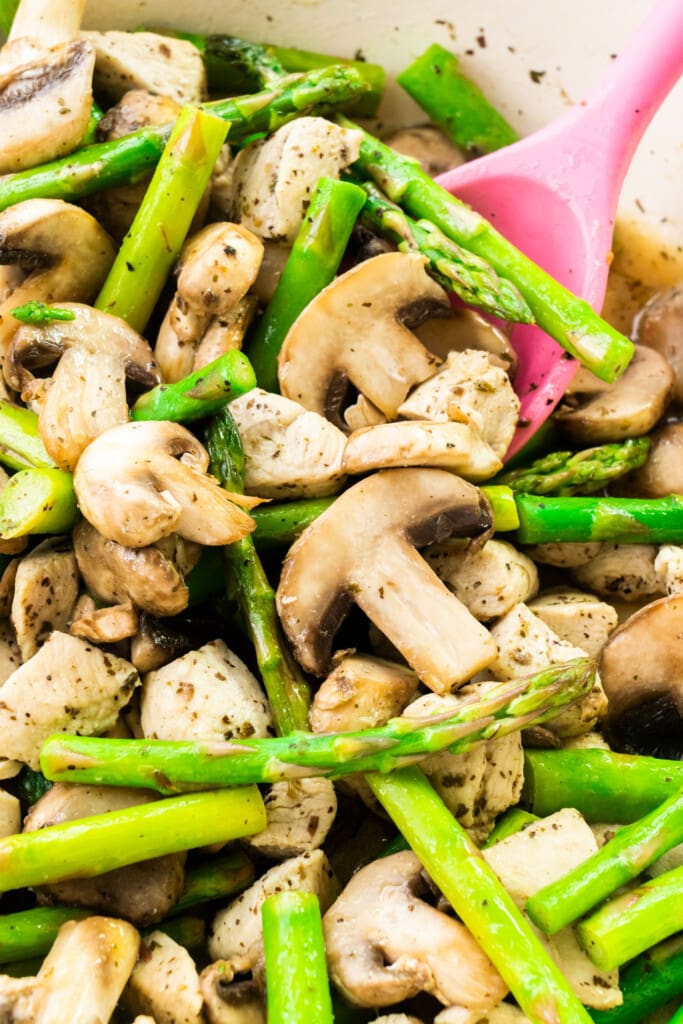 pink spoon stirring chicken, asparagus and mushrooms in skillet.