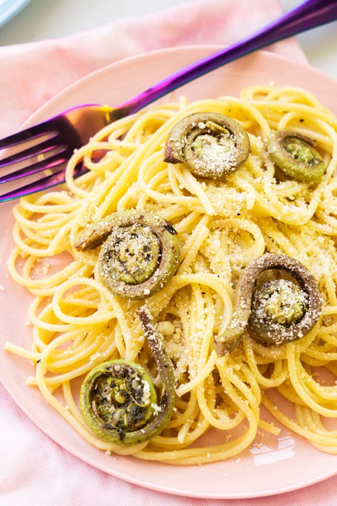 fiddleheads and pasta on pink plate with parmesan cheese on top.