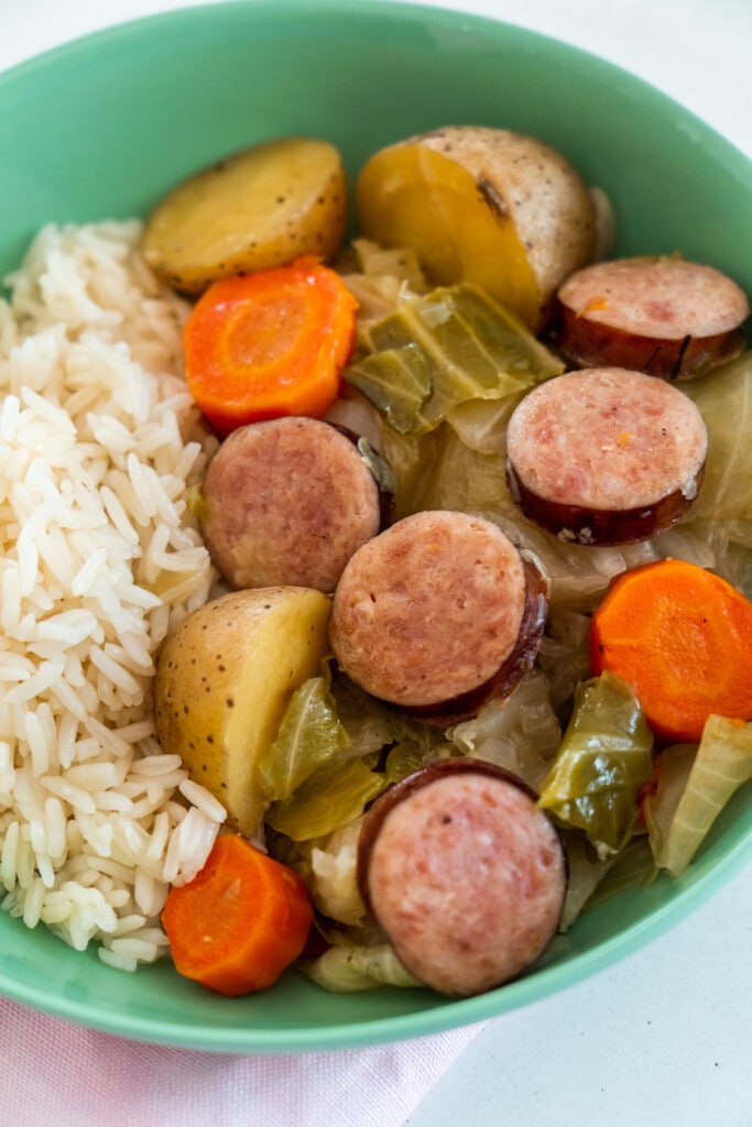 kielbasa, cabbage, carrots and potatoes next to rice in green bowl. 