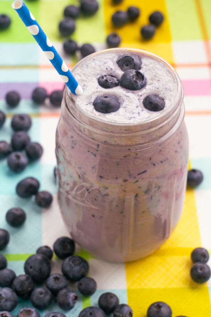 blueberry smoothie in mason jar cup on table surrounded by blueberries on table.