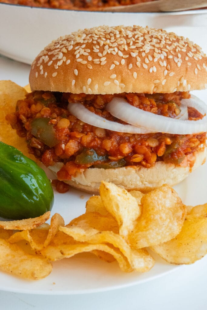 lentil sloppy joes on hamburger bun next to bbq chips and pickles on plate.