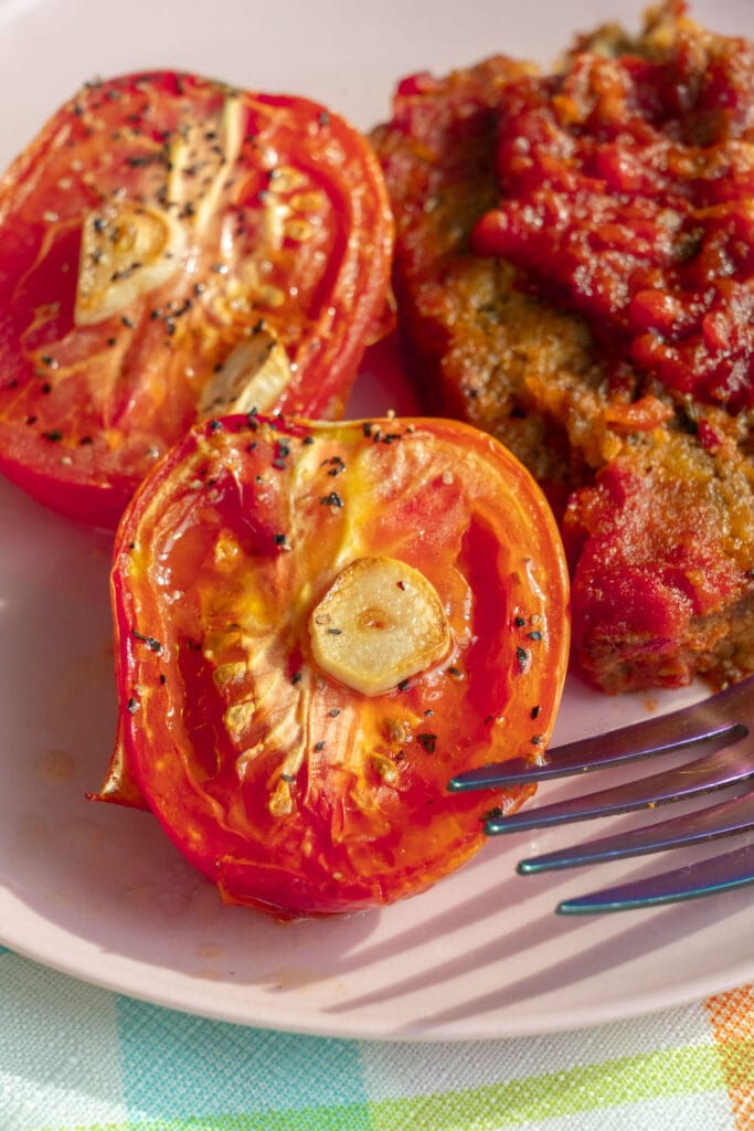 closeup of roasted roma tomato with garlic and olive oil, next to meatloaf on plate.