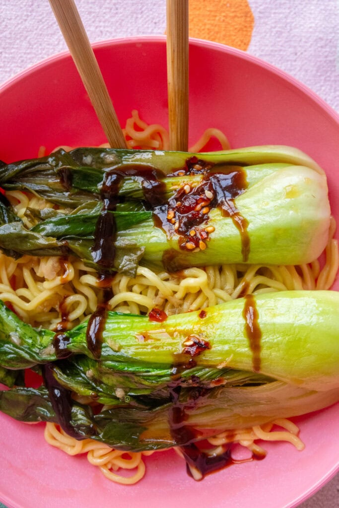 bok choy with hoisin sauce and chili sauce on top of noodles in pink bowl. 