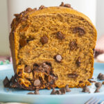 A close-up of sliced chocolate chip bread made from a bread machine rests on a blue plate. The rich brown loaf reveals swirls and chunks throughout, with extra chocolate chips scattered around the slice for added indulgence.