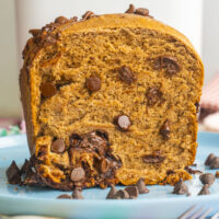 A close-up of sliced chocolate chip bread made from a bread machine rests on a blue plate. The rich brown loaf reveals swirls and chunks throughout, with extra chocolate chips scattered around the slice for added indulgence.