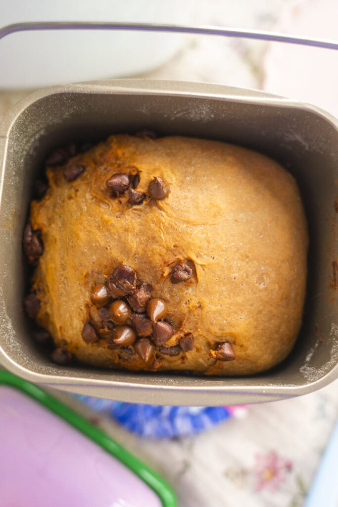 A loaf of bread machine chocolate chip bread rests in a rectangular tin, its golden-brown crust adorned with partially melted chips. The tin sits on a light-colored surface, showcasing the perfect blend of technology and taste.