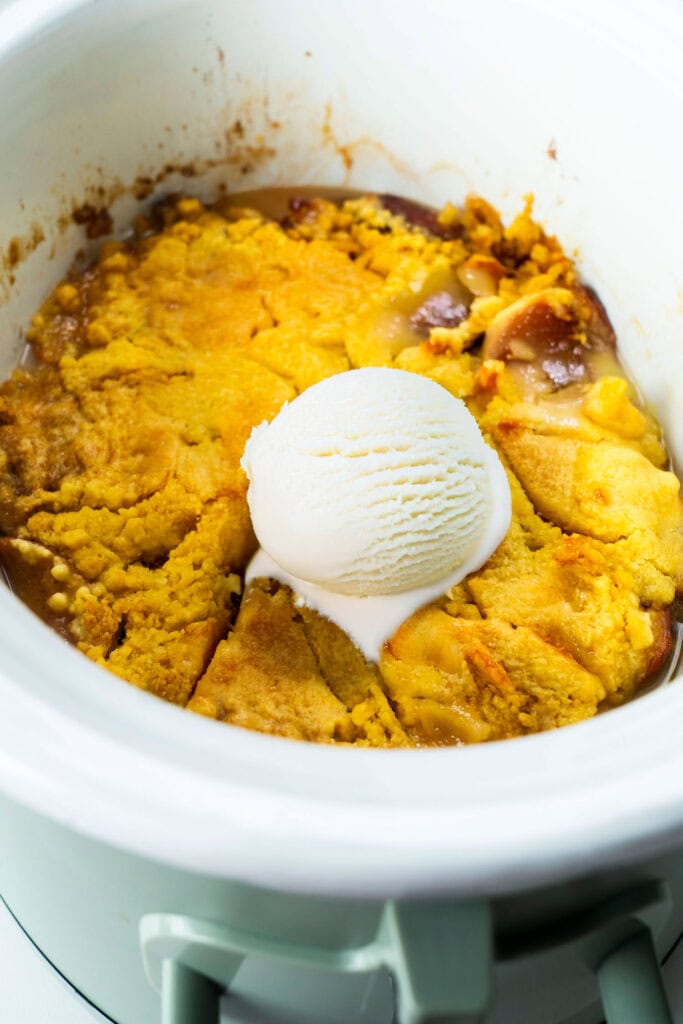 Freshly cooked peach cobbler in a white crockpot with a golden yellow cake mix topping and a scoop of melting vanilla ice cream in the center.