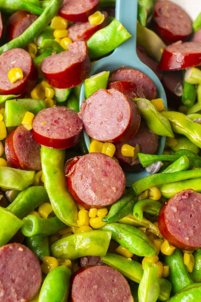 closeup of kielbasa, green beans and corn in skillet with spoon serving.