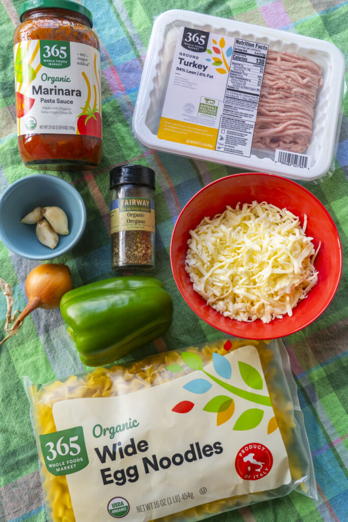 Ingredients for Cheesy Ground Turkey Noodle Casserole arranged on a green plaid tablecloth, including a package of ground turkey, a bag of wide egg noodles, marinara sauce, shredded mozzarella cheese, a green bell pepper, onion, garlic, and dried oregano.