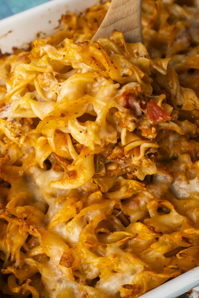 A wooden spoon scooping a serving of Cheesy Ground Turkey Noodle Casserole from a baking dish, showing melted mozzarella cheese strings, wide egg noodles, and savory meat sauce.