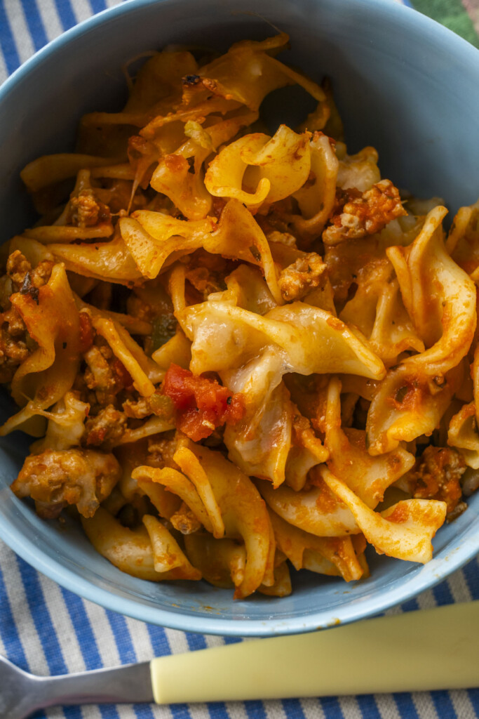 A blue bowl filled with a serving of Cheesy Ground Turkey Noodle Casserole, placed on a blue and white striped napkin with a yellow spoon.