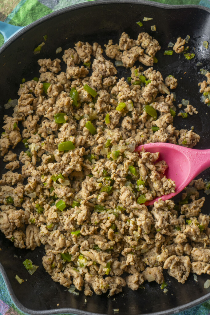 Browned ground turkey mixed with diced green bell peppers and onions cooking in a large skillet, being stirred with a pink spoon.