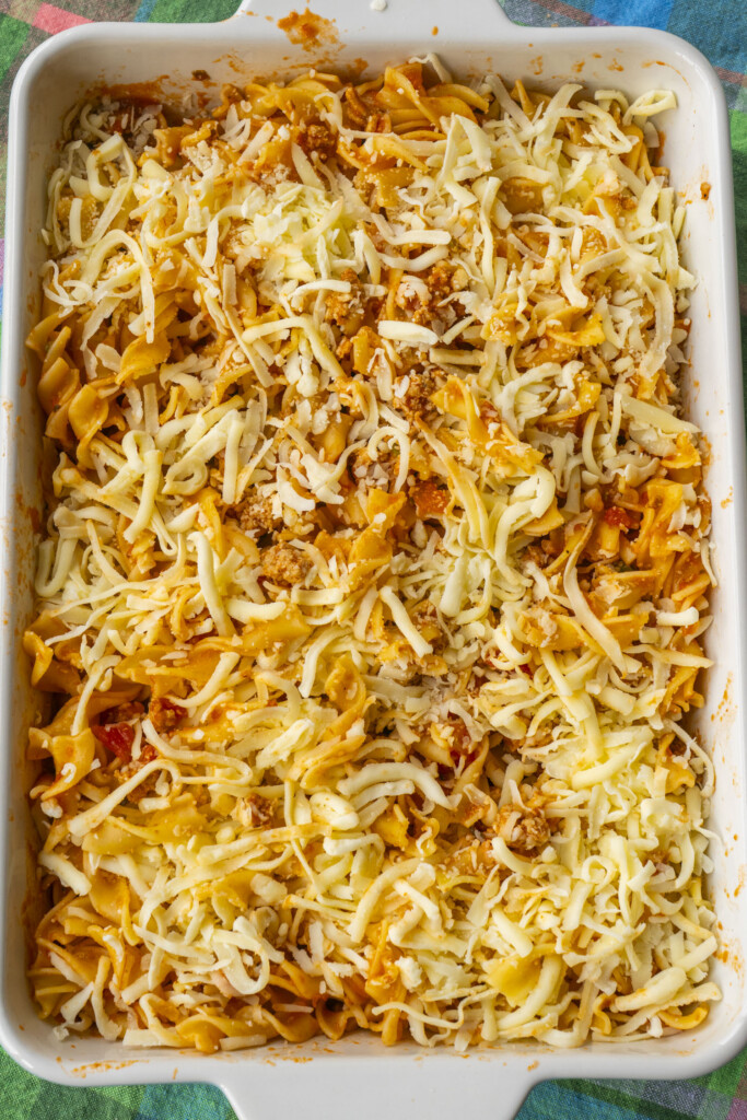 Shredded mozzarella cheese sprinkled generously over the unbaked egg noodle and turkey casserole in a white baking dish.