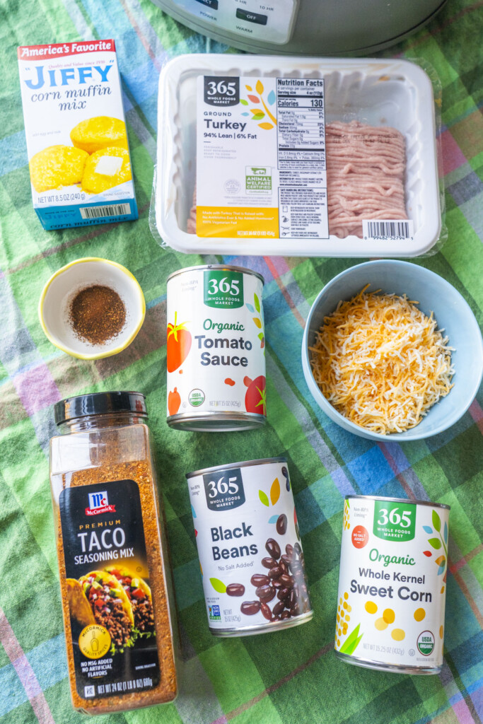 Ingredients for Crockpot Enchilada Casserole on a green plaid tablecloth, including ground turkey, Jiffy corn muffin mix, tomato sauce, black beans, corn, taco seasoning, and shredded cheese.